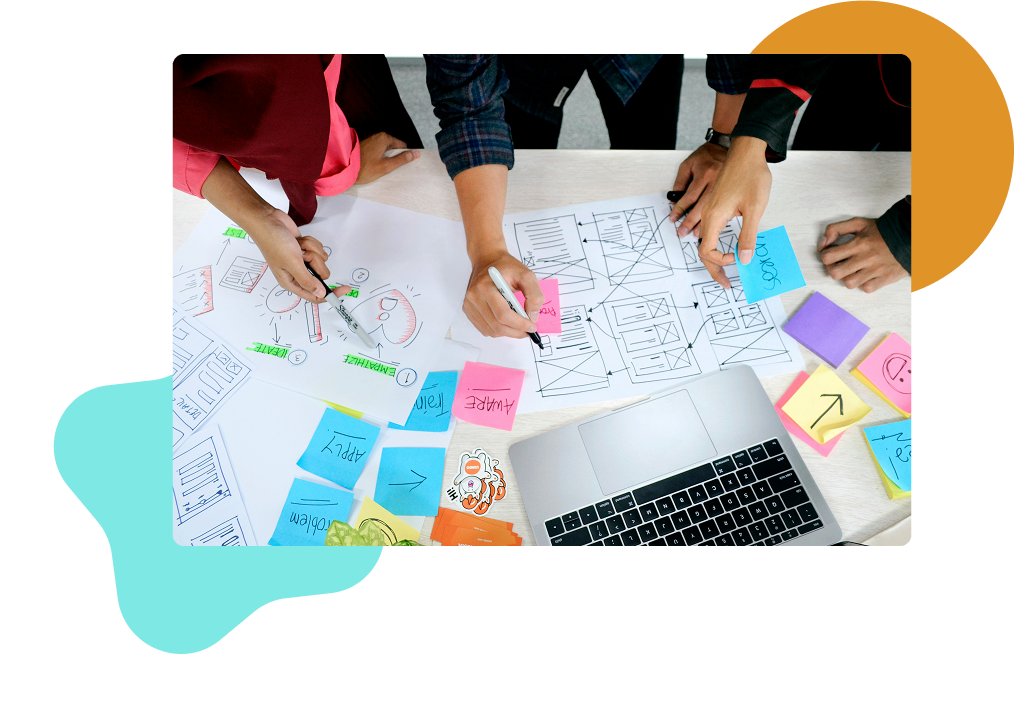 Overview of three people working on a table top with a laptop, paper, and sticky notes with strategy work on them.